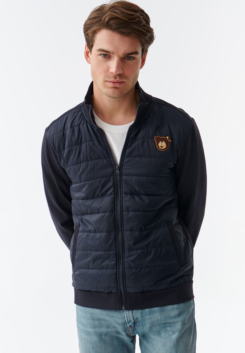 Mørkeblå zip-up jakke med kviltet front, ensfarvede ærmer og et bjørne patch på brystet. Har sidelommer og ribbede manchetter og kant.