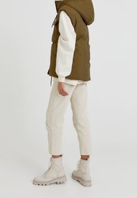 Olivgrüne Steppweste mit Kapuze über einem weißen Sweater, kombiniert mit cremefarbenen Cordhosen und hellbeigen Schnürstiefeln.