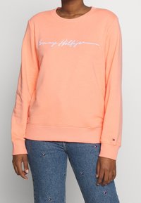 Perzikkleurig sweatshirt met geribbelde manchetten en zoom, met een wit geborduurd "Tommy Hilfiger" logo over de borst, gedragen over een blauwe spijkerbroek.