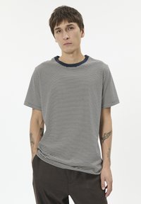 Dryžuotas marškinėlių su j�ūrinės ir baltos spalvos juostomis, su apvaliu kaklu ir trumpomis rankovėmis. Medžiaga atrodo minkšta; laisvas kirpimas, matomi tatuiruotės ant dilbio.