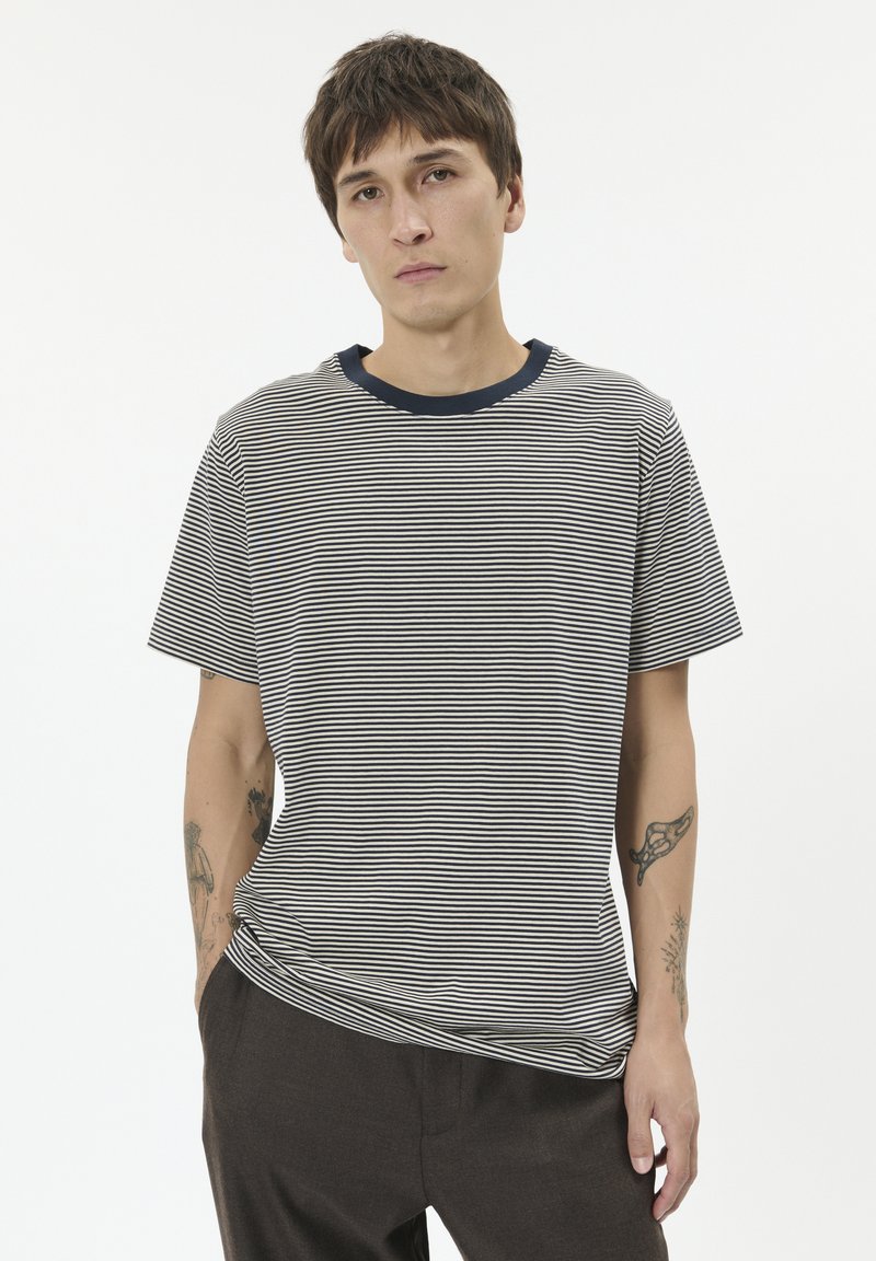 Dryžuotas marškinėlių su jūrinės ir baltos spalvos juostomis, su apvaliu kaklu ir trumpomis rankovėmis. Medžiaga atrodo minkšta; laisvas kirpimas, matomi tatuiruotės ant dilbio.