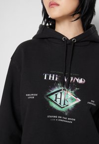 Černá mikina s kapucí a stahovací šňůrkou. Obsahuje barevný grafický design s textem, včetně "THE MIND" a "WORLDWIDE LOVER." Hladký povrch.