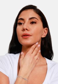 Bracciale d'argento con un delicato design a nodo indossato su una mano. Il soggetto ha capelli scuri, pelle liscia e trucco sobrio, indossando un top bianco.