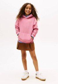 Rosa hoodie med en ficka framtill och ribbade muddar, lager-på-lager över en blå rutig skjorta, kombinerad med en brun kjol och vita sneakers.