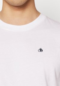 Camiseta de algodón blanca con cuello redondo y un pequeño logotipo bordado en azul marino de "&" en el lado izquierdo del pecho. Textura suave, diseño casual.