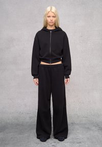 Sudadera corta negra con cremallera frontal y mangas oversized, combinada con pantalones de chándal negros de pierna ancha con cinturilla elástica.