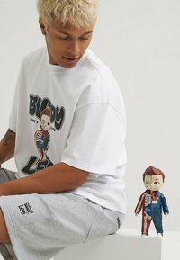 Biała, oversize koszulka z grafiką przedstawiającą postać, w połączeniu z szarymi szortami. Obok modela stoi figurka zabawki w proporcjach pół ciała.