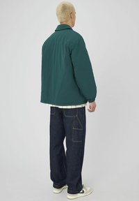 Veste teal avec une coupe décontractée, poignets côtelés et une chemise en dessous avec des rayures verticales. Pantalons cargo bleu foncé avec poches.