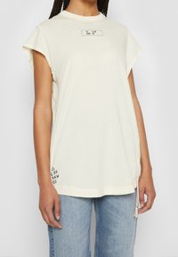 Světlé béžové tričko s krátkým rukávem, kulatým výstřihem, bočními rozparky a černým grafickým textem na přední části a dole vlevo. Měkká bavlněná tkanina.