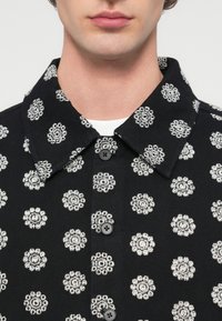 Μαύρο πουκάμισο με λουλουδένιο μοτίβο σε λευκό, με γιακά και κούμπωμα με κουμπιά. Το ύφασμα έχει υφή με ανάγλυφη εμφάνιση.