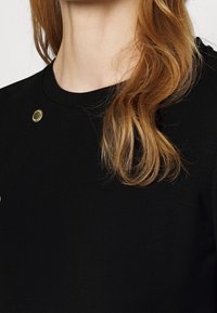 Černý oděv s kulatým výstřihem, zdobený zlatými prvky a hladkým povrchem. Vlasy splývají vedle látky.