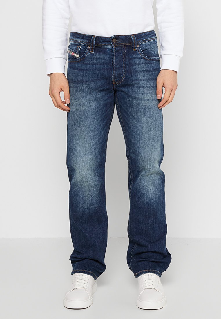 Jeans en denim bleu foncé avec une coupe droite, présentant un léger délavage, une fermeture à bouton et cinq poches, portés avec des baskets blanches.