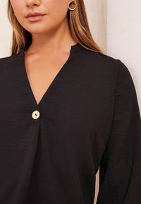 Maglietta nera con scollatura a V, caratterizzata da un dettaglio con bottone dorato, realizzata in un tessuto testurizzato con maniche lunghe e una vestibilità morbida.