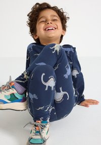 Bambino sorridente con capelli ricci seduto sul pavimento, indossa un pigiama blu navy con stampa di dinosauri e sneaker colorate con lacci.
