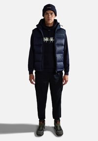 Marine gewatteerd vest over een marine hoodie met grafische print, gecombineerd met zwarte cargo-broeken en grijs-groene sneakers. Accessoires omvatten een marine beanie.