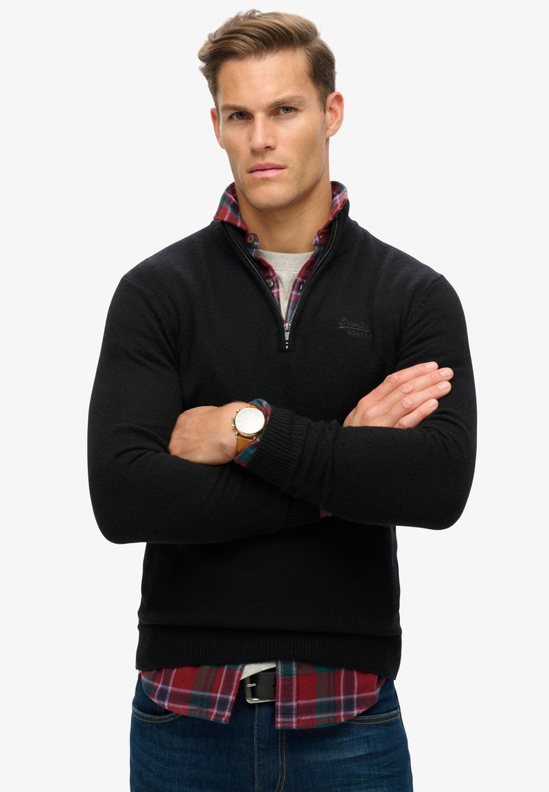 Maglione zip-up nero realizzato in tessuto a maglia testurizzato, indossato sopra una camicia a quadri rossa, con una maglietta grigia sotto e jeans blu.
