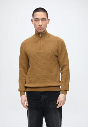 Uomo che indossa un maglione marrone a coste con mezza cerniera e jeans neri, in piedi su uno sfondo bianco uniforme.
