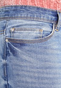 Jeans de denim azul claro con un diseño de cinco bolsillos, con parches deslavados y costuras en contraste. Se puede ver un pequeño botón decorativo.