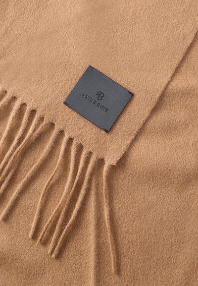 Pehmeä kamelinvärinen huivi, jossa on hapsutetut reunat ja musta neliskulmainen kiinnitetty etiketti, johon on painettu "ANINE BING" ja pyöreä logo.