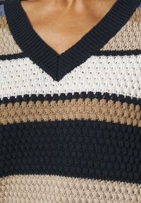 Stickad tröja med V-ringning och horisontella ränder i beige, svart och krämfärg, som visar ett strukturerat mönster.