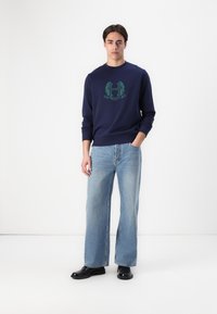 Navy blue sweatshirt with green logo, crew neckline, long sleeves, paired with light blue, wide-leg jeans and black shoes. Simple, casual outfit.
