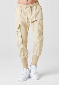 Beige cargo-housut, joissa on vetoketjukaulus, vetosäätövyötärö ja ribbireunat. Yhdistettynä valkoisiin lenkkitossuihin.