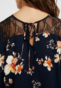 Blusa floreale con base blu navy decorata con fiori arancioni e bianchi, inserto in pizzo nero, e chiusura con nastro sul retro del collo.