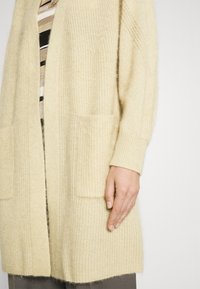 Cardigan lavorato a maglia beige con vestibilità ampia, caratterizzato da una trama a coste, apertura frontale e due tasche anteriori. Le maniche lunghe superano il polso.