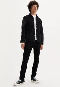 Melna džinsa jaka ar spiedpogām, kombinēta ar baltu T-kreklu, melnām džinsu biksēm un tumšām sporta kurpēm. Ikdienišķs siluets ar redzamām šuvju detaļām.