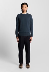 Mörkblå stickad tröja med ribbade manschetter, tillsammans med svarta byxor och bruna mönstrade sneakers. Modell som står framför en ljus bakgrund.