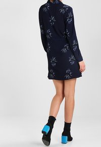 Abito navy con stampa floreale in tonalità di blu e verde. Presenta maniche lunghe e una vita aderente. Indossato con stivali neri alla caviglia e tacchi blu.