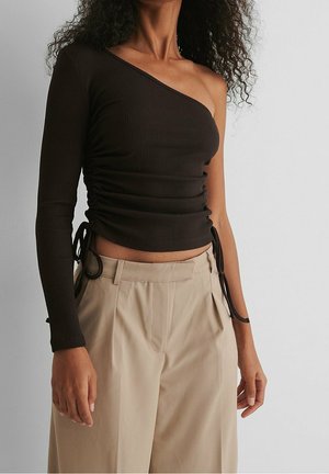 Kvinde iført en sort ribbet, asymmetrisk bluse med én lang ærme, sidebind og højtaljede, beige bukser med vide ben mod en grå baggrund.