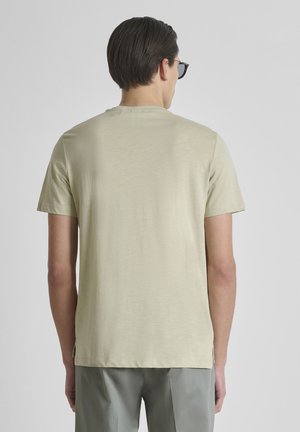 Hombre con camiseta de manga corta beige y pantalones grises, visto desde atrás sobre un fondo liso de color gris claro.