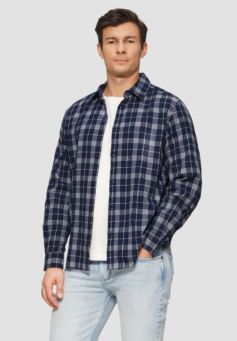 Navyblauwe geruite shirt met witte accenten, button-down kraag, lange mouwen en een ontspannen pasvorm, gedragen over een wit T-shirt en lichtblauwe jeans.