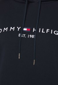 Tmavě modrá mikina s kapucí z bavlněné směsi, s bílým vyšitým názvem značky s červenými a modrými akcenty a šňůrkami s pozlacenými konci.