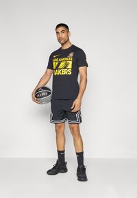Camiseta atlética negra con texto amarillo "Los Angeles Lakers", combinada con pantalones cortos de malla negros con ribete blanco y zapatos deportivos negros.