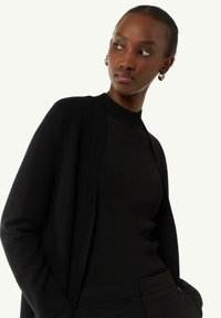 Schwarze Strickjacke mit geripptem Muster, offener Front und langen Ärmeln, die über einem schwarzen gerippten Top und kombiniert mit schwarzen Hosen getragen wird.