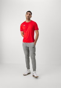 Camiseta deportiva roja de manga corta con logo blanco, combinada con pantalones de chándal grises con un logo negro y zapatillas blancas con suelas marrones.