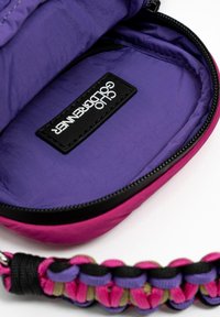 Compact tas met een gebogen vorm. De buitenkant is van roze stof; de binnenkant heeft een paarse voering. Zwart rits en label; gevlochten handvat in meerdere kleuren.