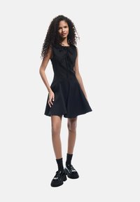 Schwarzes ärmelloses Kleid mit plissiertem Rock und einer Frontbindedetail. Das Modell trägt schwarze Socken und glänzende Plateauschuhe. Glatte Stofftextur.