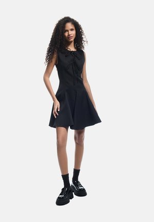 Robe noire sans manches avec une jupe plissée et un détail à nouer à l'avant. Le modèle porte des chaussettes noires et des chaussures à plateforme brillantes. Texture de tissu lisse.