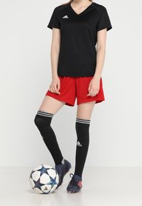 Jersey negro de manga corta, pantalones cortos rojos, calcetas negras hasta la rodilla con rayas blancas y zapatillas deportivas negras, de pie al lado de un balón de fútbol.