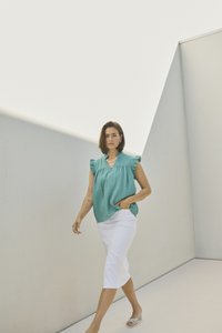 Haut turquoise à volants en tissu texturé, associé à une jupe blanche longueur genou et des sandales légères, sur fond minimaliste.