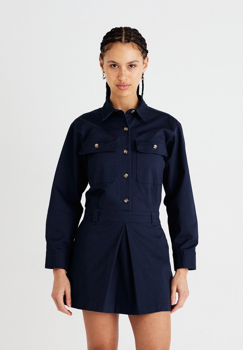 Rochie camasa bleumarin cu maneci lungi, doua buzunare pe piept, nasturi metalici si fusta plisata, confectionata dintr-un material din bumbac.