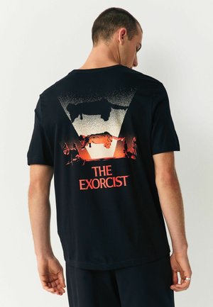 Schwarzes Baumwoll-T-Shirt mit einem grafischen Druck auf der Rückseite, der rote und weiße Elemente enthält und eine Szene aus "Der Exorzist" darstellt.