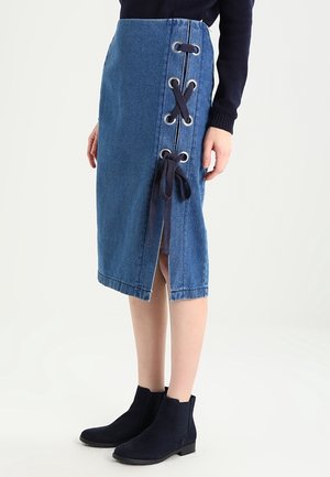 Woman wearing a knee-length blue denim skirt with side lace-up detail and black ankle boots, standing against a plain white background.