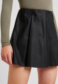 Jupe en cuir plissée noire avec une texture lisse. Fournie avec une taille ajustée et des détails de coutures subtils pour un intérêt supplémentaire.