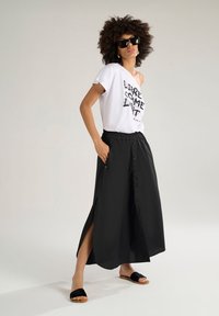 T-shirt bianca con grafica "LIBRE COME LMT" abbinata a una gonna nera alla caviglia con spacchi laterali e dettagli di bottoni. Sandali neri.