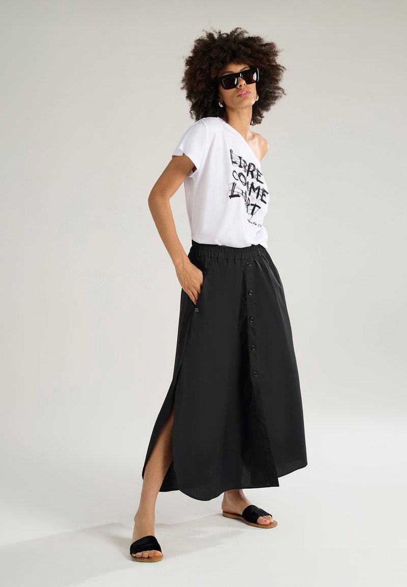 T-shirt bianca con grafica "LIBRE COME LMT" abbinata a una gonna nera alla caviglia con spacchi laterali e dettagli di bottoni. Sandali neri.