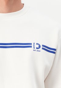 White sweatshirt featuring blue horizontal stripes and text "D" and "EST. 2007," with ribbed crew neckline and soft texture.